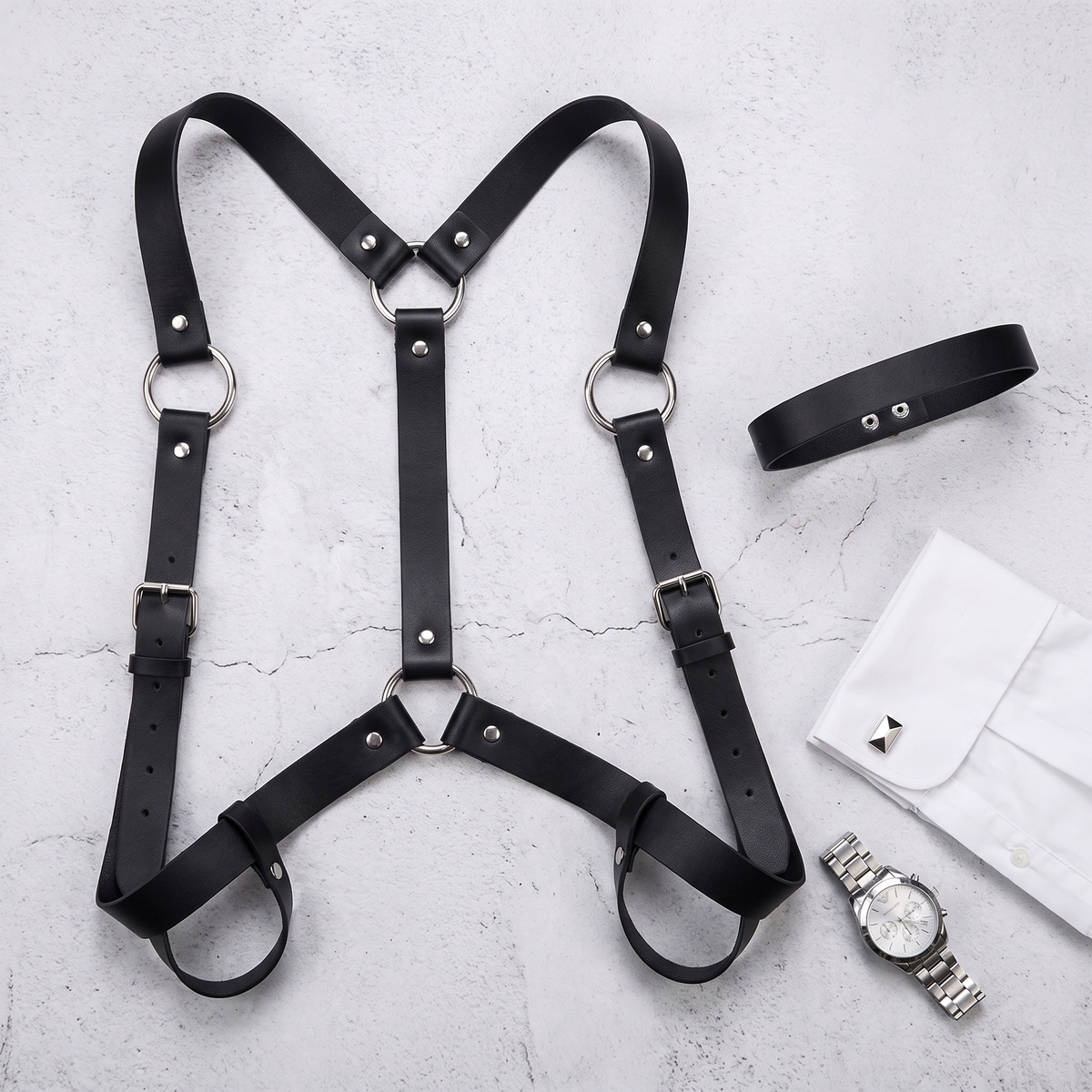 Black faux leather chest harness laid flat on concrete beside a shirt cuff and a watch