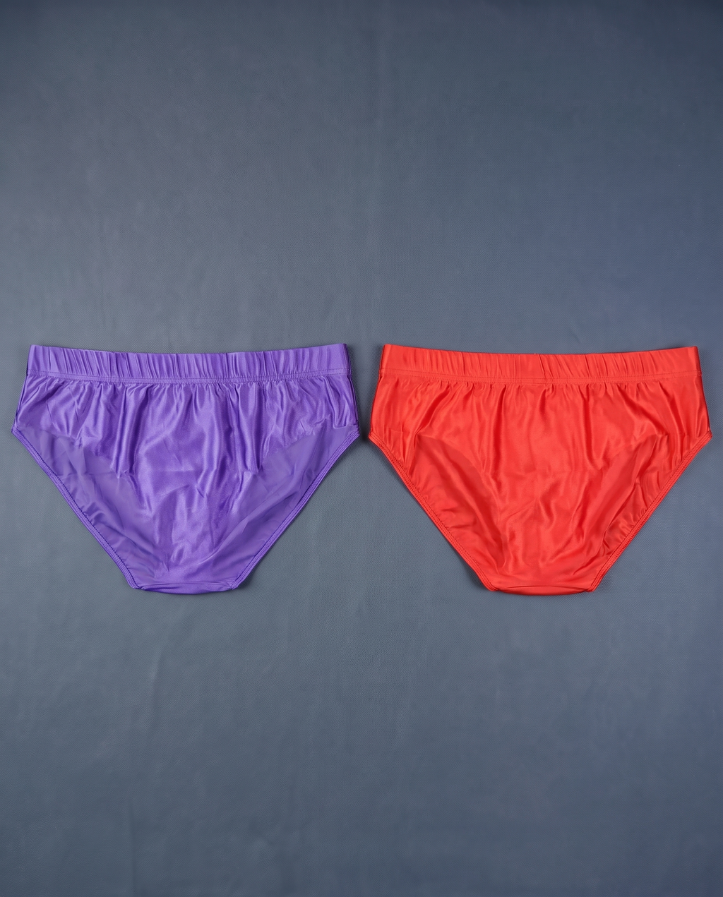 Back view of purple and red men's satin-finish briefs laid flat on slate
