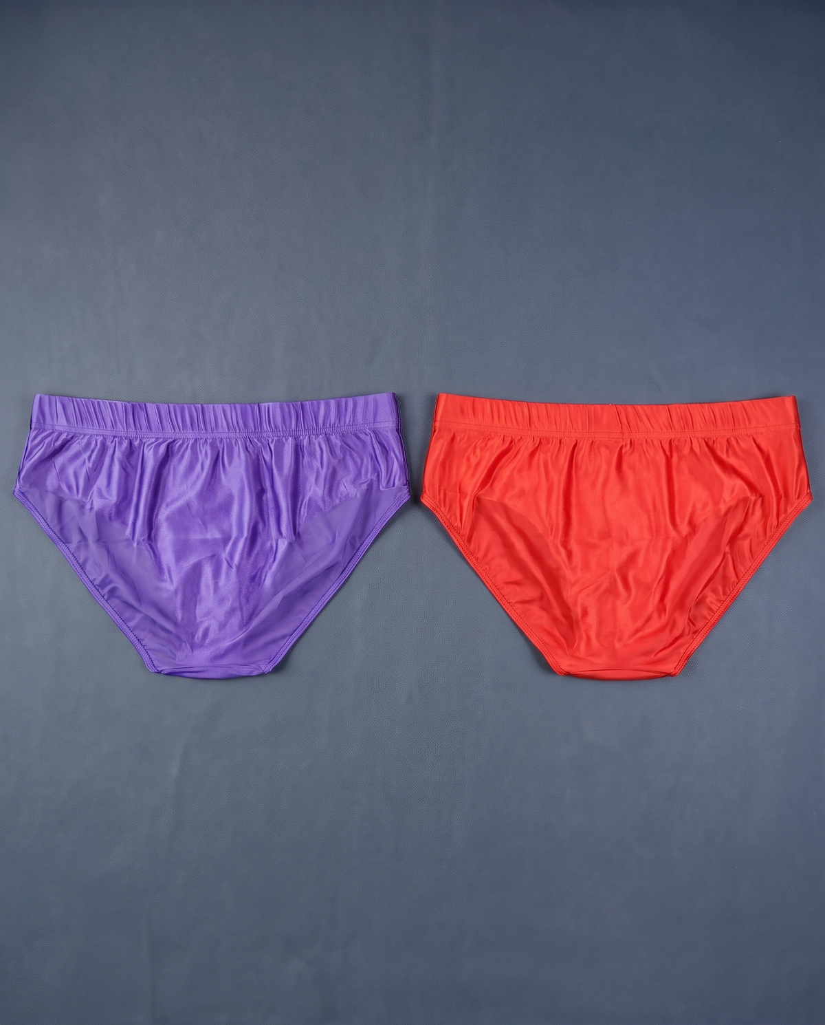 Back view of purple and red men's satin-finish briefs laid flat on slate